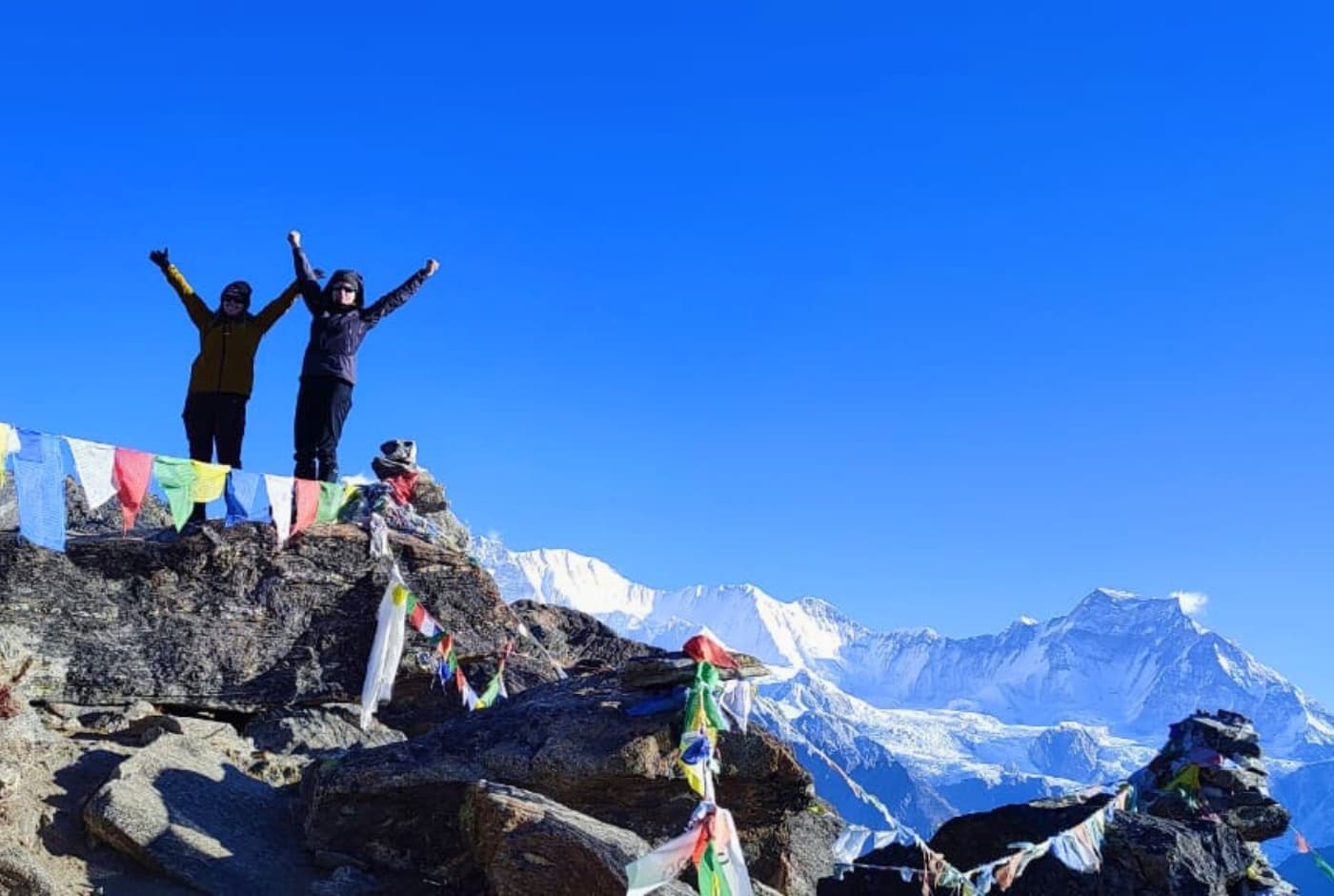 Two hikers celebrating at the summit with prayer flags and snow Capped mountains