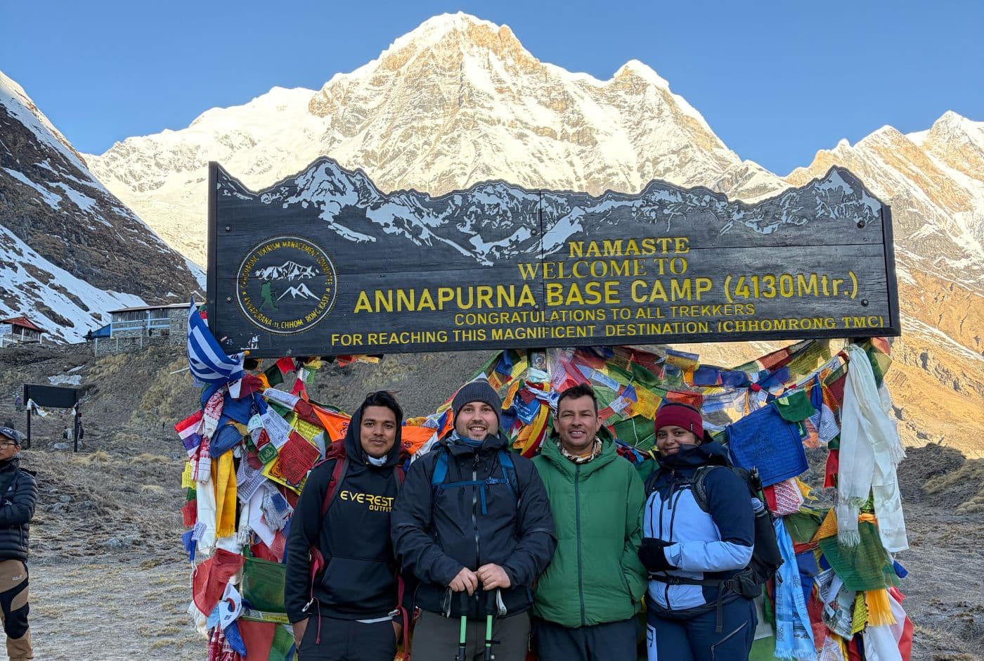 Group of trekkers standing at Annapurna Base Camp (4,130m) beneath the welcome sign, with colorful prayer flags and Annapurna South peak glowing in morning light