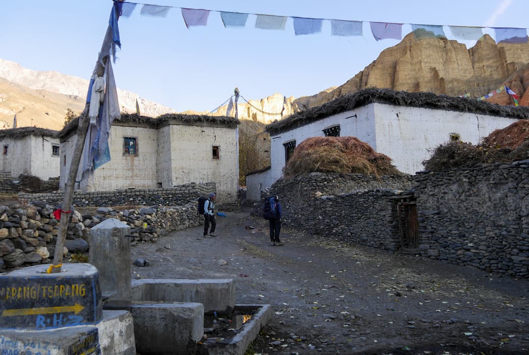 Trekkers Walking Through Charang (Tsarang) Village With Prayer Flags And Traditional White Stone Houses On The Upper Mustang Trek Nepal