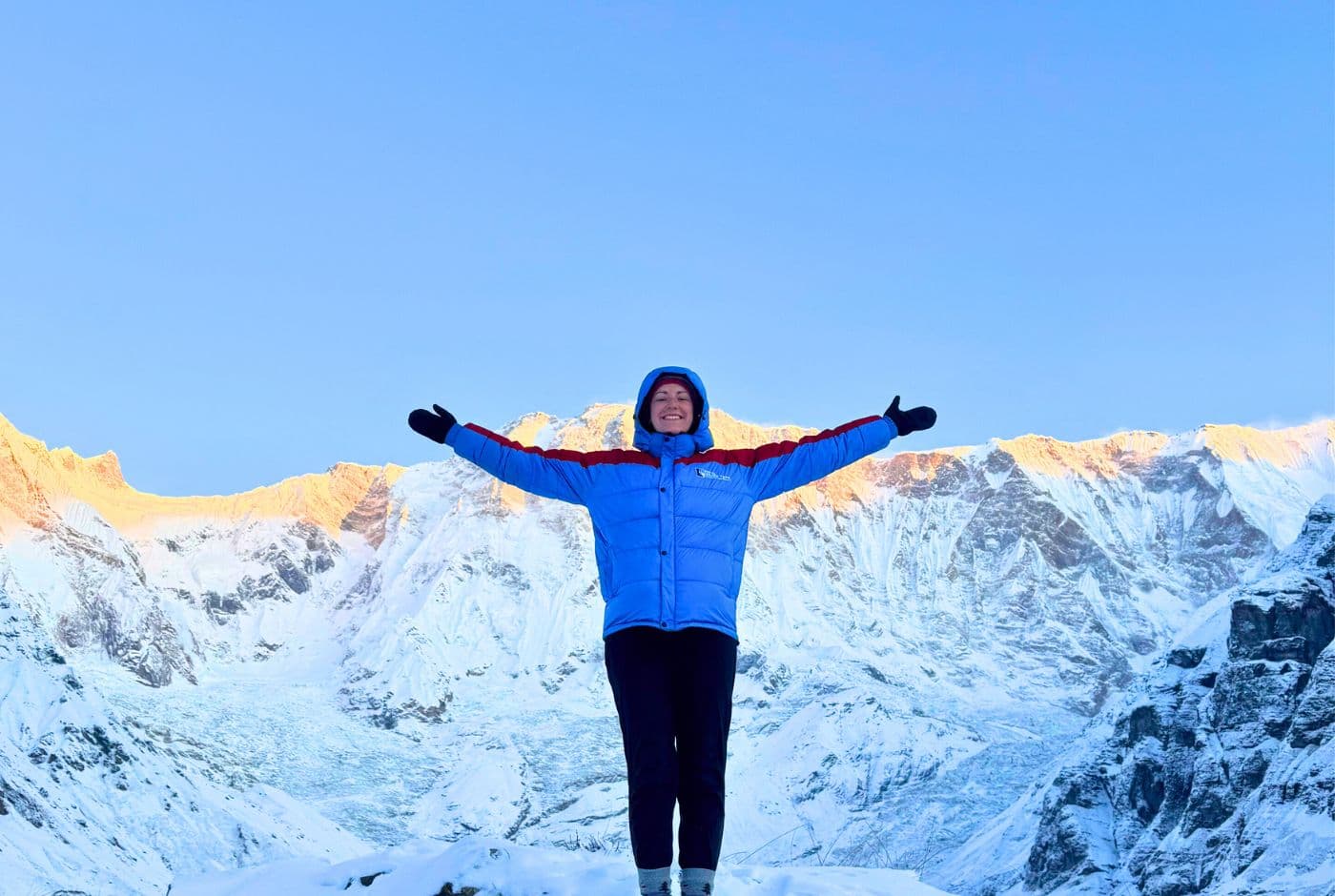 Trekker with arms outstretched at Annapurna Base Camp on ABC Trek, Annapurna I in back amid snowy peaks and blue skies.