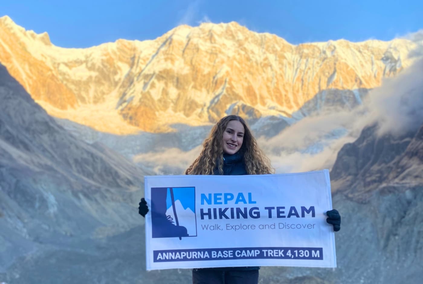 Female trekker holding Nepal Hiking Team banner at Annapurna Base Camp, Annapurna I glowing behind