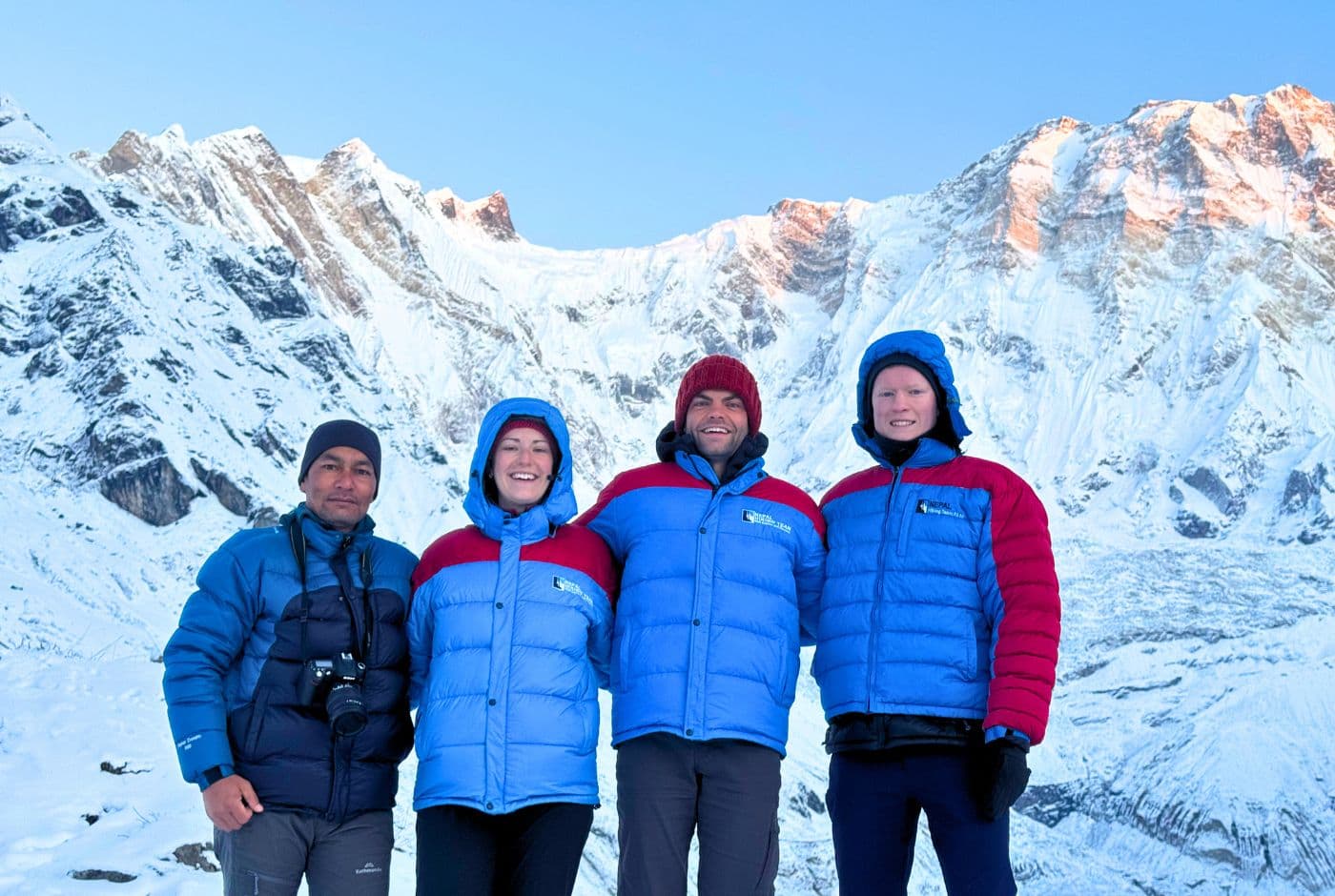 Four trekkers posing at Annapurna Base Camp (4,130m) during the Annapurna Base Camp Trek, with Annapurna I rising on the right inside the snowy Annapurna Sanctuary, Nepal
